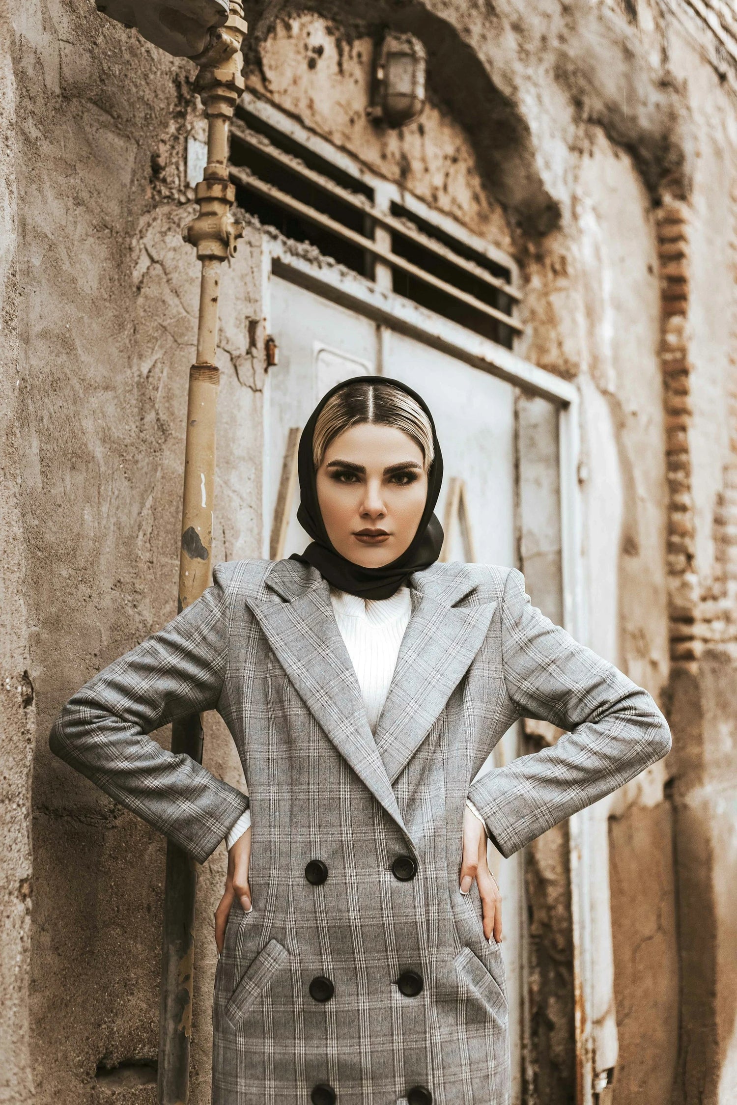 Donna vestita con giubbotto lungo, maglioncino e foulard in testa, con le braccia appoggiate sui fianchi, in contesto cittadino di un quartiere storico