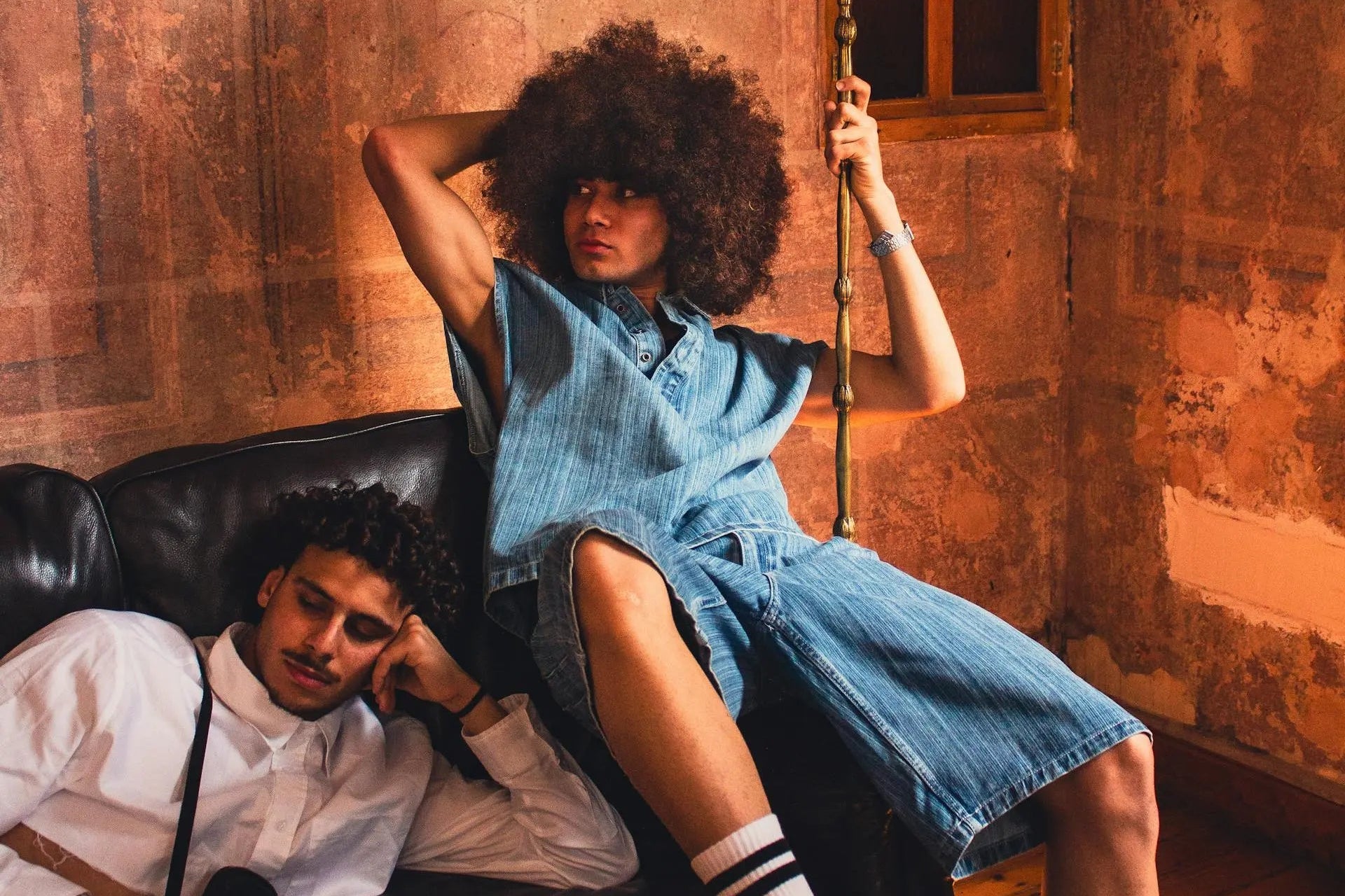 Ragazzo afro in posa su divano, con un ragazzo elegante con camicia bianca di fianco, sempre in posa