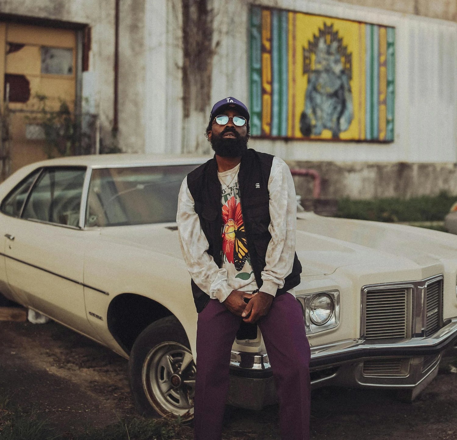 Uomo vestito streetwear appoggiato a una macchina vintage anni 90, con sfondo murales da città caraibica